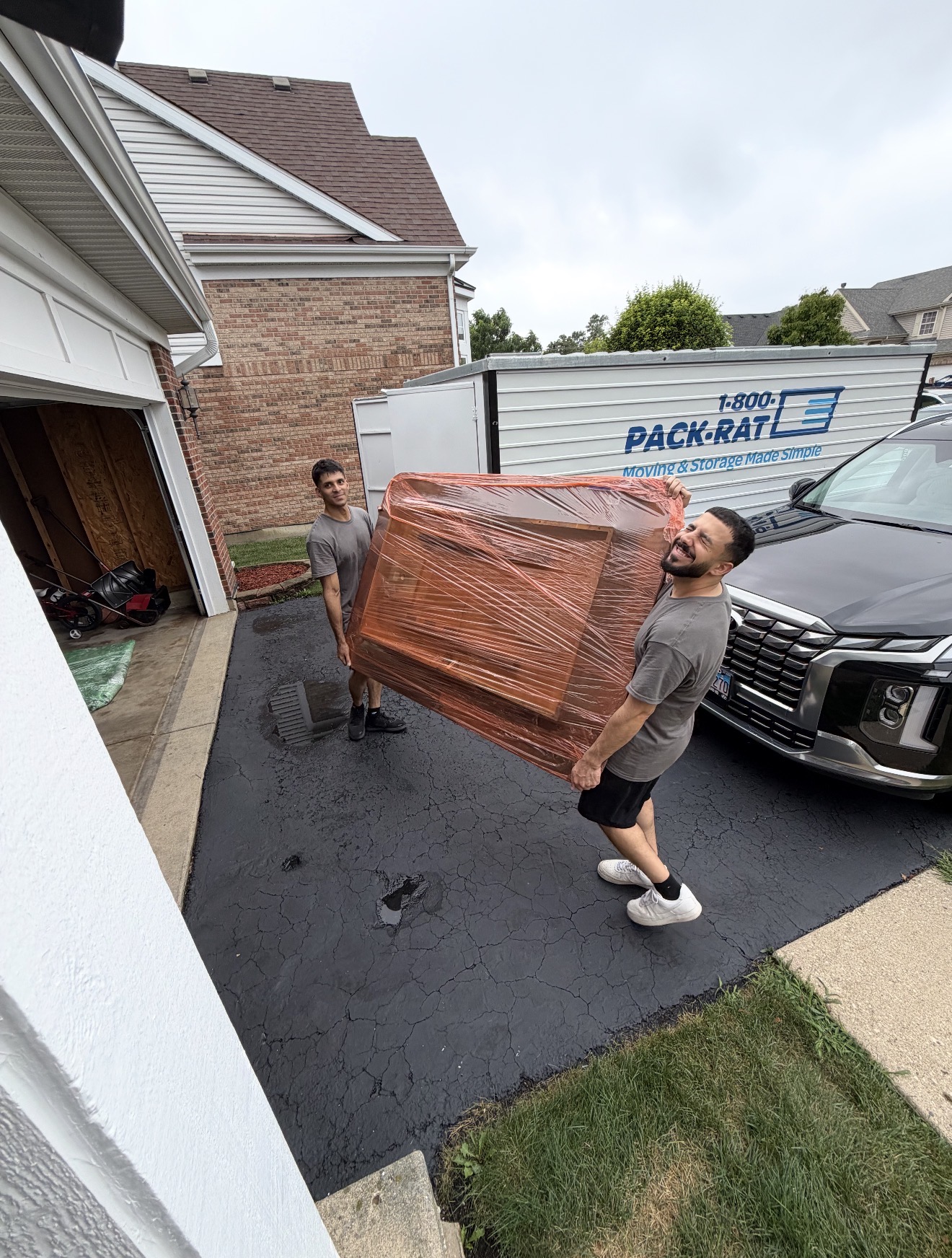 Two movers carrying a shrink-wrapped piece of furniture outside a home with a Pack-Rat container